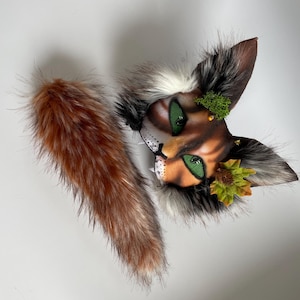 May include: A handcrafted cat mask with a faux fur tail. The mask features a detailed painted face with green eyes, brown and orange fur, and decorative moss and leaves. The tail is a mix of brown and orange faux fur.
