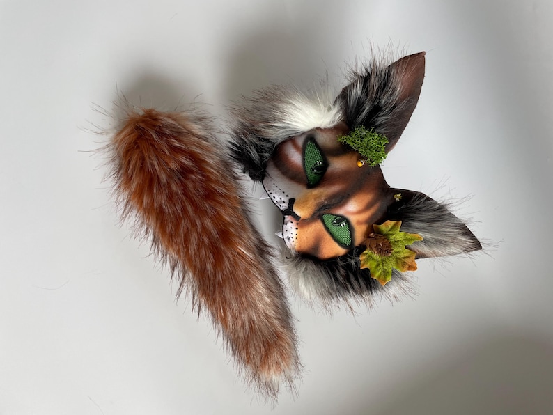 May include: A handcrafted cat mask with a faux fur tail. The mask features a detailed painted face with green eyes, brown and orange fur, and decorative moss and leaves. The tail is a mix of brown and orange faux fur.