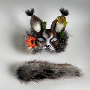 May include: A handcrafted lynx mask and tail set. The mask has a painted face with white, brown, and black fur details, decorated with artificial flowers and foliage. The tail is made of soft, grey and brown faux fur.