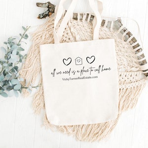 May include: A white canvas tote bag with black text that reads "all we need is a place to call home" and a small house icon with two hearts on either side. The text "VickyTurnerRealEstate.com" is printed below the main text.