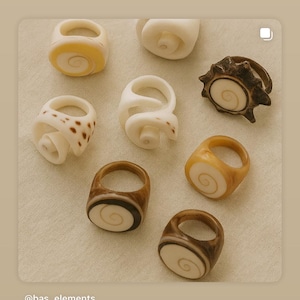 Op de afbeelding: Een verzameling handgemaakte ringen in verschillende ontwerpen en kleuren. De ringen hebben een spiraalvormig ontwerp, sommige in wit, bruin en gele tinten. De ringen worden op een neutrale achtergrond gepresenteerd. De tekst "@bas_elements" is zichtbaar.