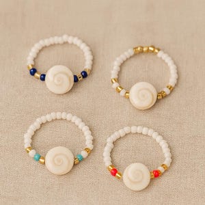 May include: Four beaded rings with a central, cream-colored spiral design. The rings are white with accents of blue, gold, turquoise, and red beads. The rings are displayed on a neutral, textured surface.