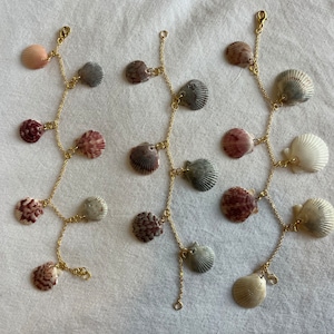 May include: Three seashell garlands with gold-colored chains and clasps. The seashells are in various colors, including pink, gray, and white, and are arranged in a cascading pattern. The garlands are displayed on a white surface.