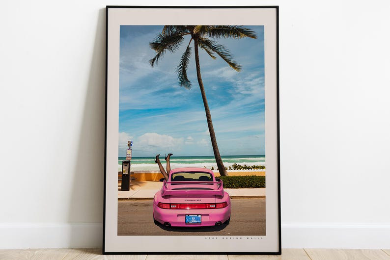 Puede incluir: Un Porsche 911 rosa con el techo bajado estacionado en una playa con una palmera de fondo. El coche tiene una matr&iacute;cula que dice "ONE CRAZY LIFE".
