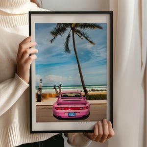Puede incluir: Un Porsche 911 rosa estacionado en una playa con una palmera de fondo. Las piernas de una persona sobresalen de la parte trasera del coche.