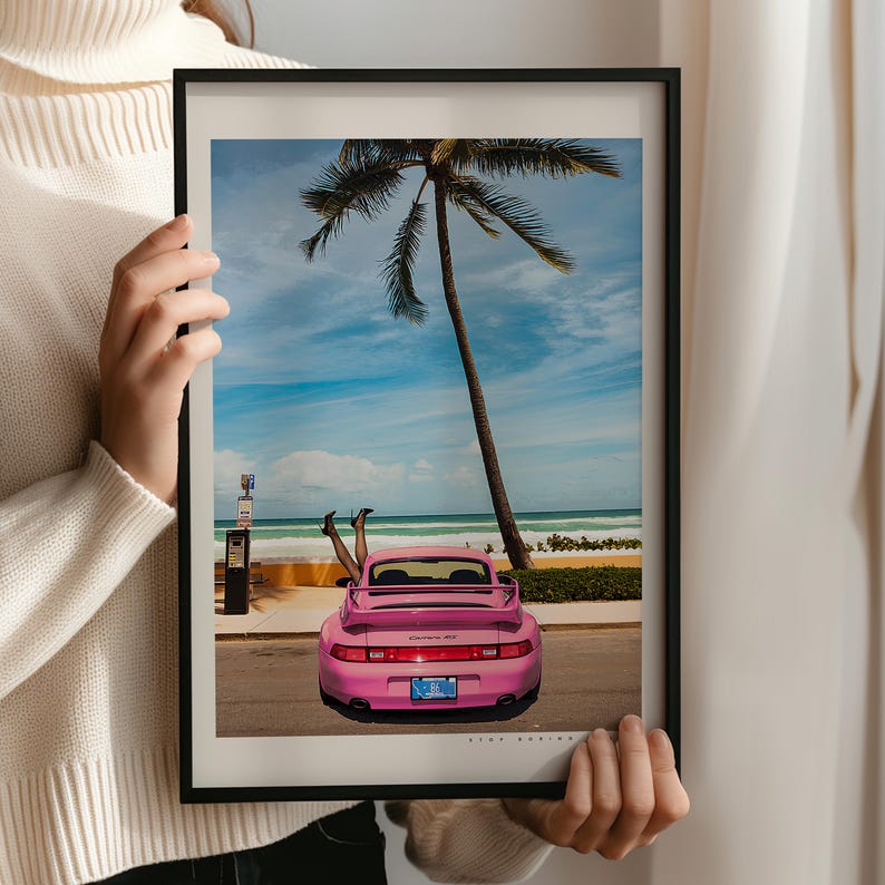 Puede incluir: Un Porsche 911 rosa estacionado en una playa con una palmera de fondo. Las piernas de una persona sobresalen de la parte trasera del coche.