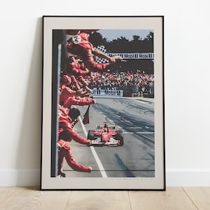 K&ouml;nnte beinhalten: Ein roter Formel-1-Rennwagen rast &uuml;ber die Ziellinie, im Hintergrund jubelt eine Menschenmenge in roten Overalls. Das Auto ist ein Ferrari mit der Nummer 1 an der Seite. Die Worte "Mobil 1" sind auf der Strecke zu sehen.