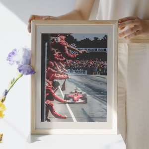 K&ouml;nnte beinhalten: Ein gerahmtes Foto eines roten Rennwagens, der an einer Menge von Menschen vorbeif&auml;hrt, die rote Flaggen schwenken. Das Auto ist mit "Marlboro" beschriftet und im Hintergrund befindet sich ein Schild mit der Aufschrift "Mobil 1".