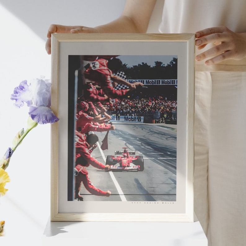 K&ouml;nnte beinhalten: Ein gerahmtes Foto eines roten Rennwagens, der an einer Menge von Menschen vorbeif&auml;hrt, die rote Flaggen schwenken. Das Auto ist mit "Marlboro" beschriftet und im Hintergrund befindet sich ein Schild mit der Aufschrift "Mobil 1".