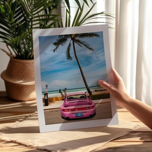 Puede incluir: Un Porsche 911 rosa estacionado en una carretera de playa con una palmera de fondo. El coche tiene una matr&iacute;cula que dice "The-Design-Studio".
