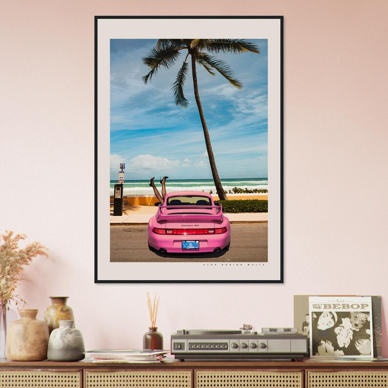 Puede incluir: Un Porsche 911 rosa con las piernas de una mujer asomando por la ventana trasera. El coche est&aacute; aparcado en una calle con una palmera de fondo. La playa se ve en la distancia.