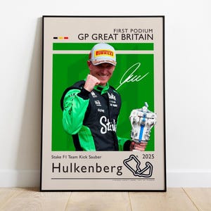 以下が含まれることがあります： 緑と黒のレーシングスーツを着たF1ドライバーがトロフィーを持っている額入りのポスター。ポスターには「GP GREAT BRITAIN」と「Hulkenberg 2025」の文字が入っています。ドライバーはピレリのロゴが入ったキャップをかぶっています。