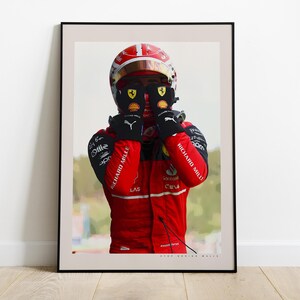 May include: Framed print of a Formula 1 driver in a red racing suit and helmet. The driver is wearing black gloves and is holding them up to his face. The print is in a black frame and is displayed on a wooden floor.