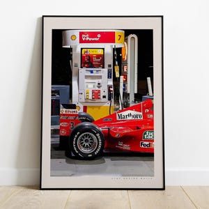 May include: A red Formula One race car with the words "Marlboro" and "Tic Tac" on the side, parked at a petrol station with a Shell V-Power sign. The car has a large tyre in the foreground.