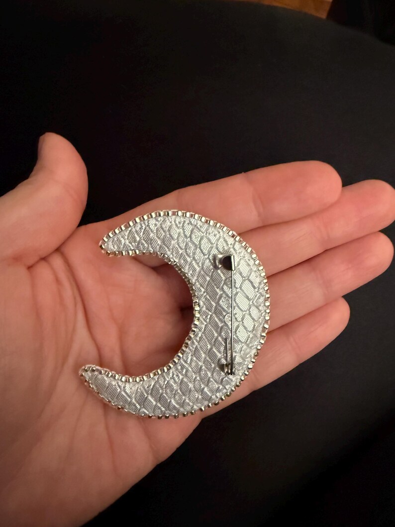 May include: A silver crescent moon brooch with a textured, snake-skin like design. The brooch has a silver pin and catch on the back.