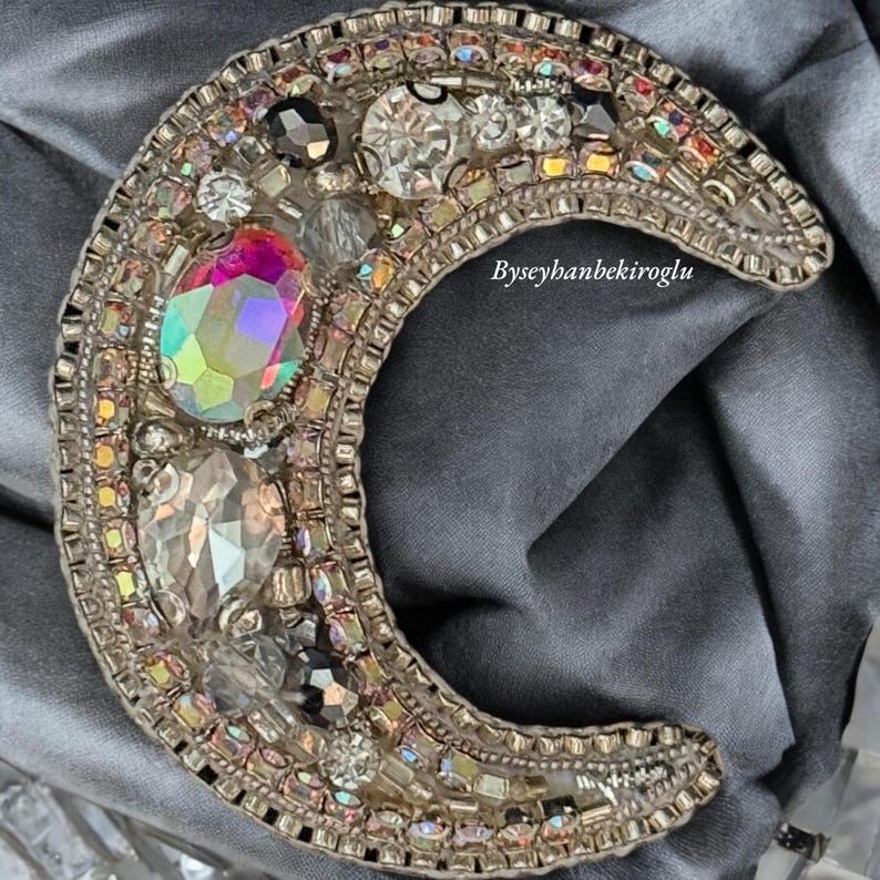 May include: A crescent moon-shaped brooch with a silver base and a variety of colourful rhinestones and beads. The brooch is decorated with a variety of colours, including pink, blue, green, and black. The brooch is on a grey fabric background.