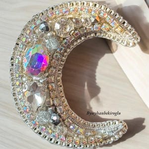 May include: A silver crescent moon brooch featuring iridescent crystals and rhinestones. The brooch is adorned with a variety of sizes and shapes of crystals, creating a sparkling effect.