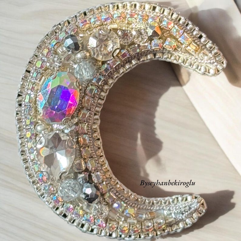 May include: A silver crescent moon brooch featuring iridescent crystals and rhinestones. The brooch is adorned with a variety of sizes and shapes of crystals, creating a sparkling effect.