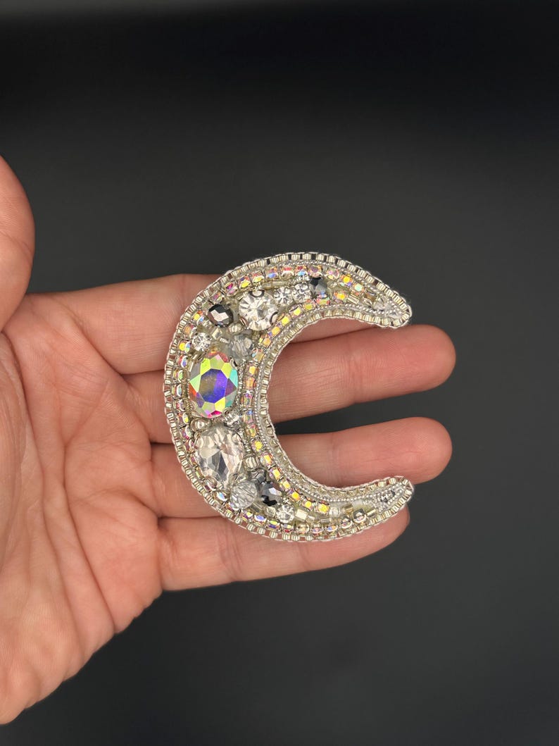 May include: A silver crescent moon brooch with iridescent and clear rhinestones. The brooch is beaded and has a silver backing.