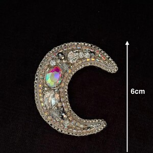 May include: A silver crescent moon-shaped patch with iridescent rhinestones and beads. The patch measures 6 cm in diameter.