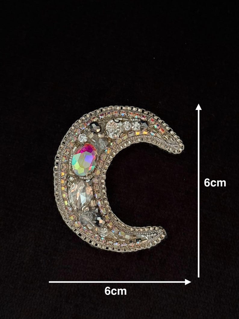 May include: A silver crescent moon-shaped patch with iridescent rhinestones and beads. The patch measures 6 cm in diameter.