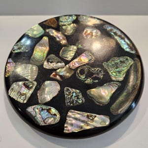 May include: Round, black decorative object inlaid with iridescent abalone shell pieces. The shell fragments vary in size and shape, displaying a range of colors including green, blue, and gold. The object has a smooth, polished surface.