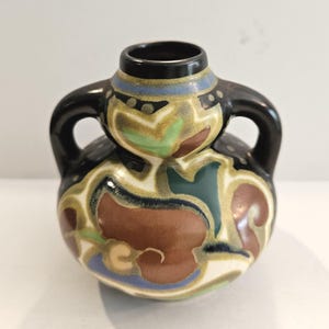 May include: A small, decorative ceramic vase with two handles. The vase features a black rim and handles, with a body adorned with a colorful floral pattern in shades of brown, green, blue, and cream. The vase is approximately 4 inches tall.