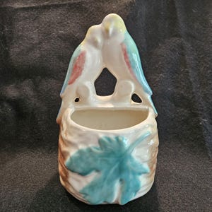 May include: A vintage ceramic wall pocket featuring two birds perched together. The birds have white, blue, and red accents. The pocket has a leaf design in blue and brown. The piece is likely for decorative use.