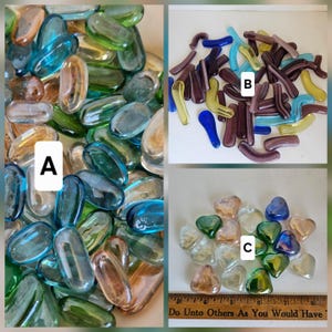 May include: A collection of colorful glass pebbles in various shapes and sizes. The pebbles are in shades of blue, green, yellow, and brown. Some are oval, others are curved, and some are heart-shaped. The text "Do Unto Others As You Would Have" is visible.