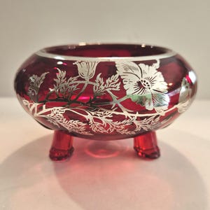 May include: A ruby red glass bowl with three small feet. The bowl features a silver floral design around the exterior. The bowl is a decorative piece, likely for display or holding small items.