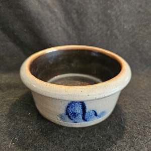 May include: A small, round stoneware bowl with a light gray exterior and a dark brown interior. The bowl features a blue floral design on the side. The rim is slightly flared, and the bowl has a rustic, handmade appearance.