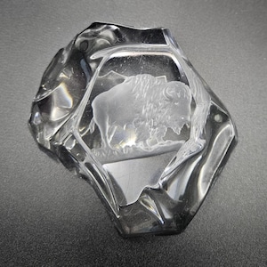 May include: A clear, irregularly shaped glass paperweight featuring an etched, frosted-white image of a bison standing in profile. The glass has a smooth, polished surface with faceted, uneven edges that create a reflective, multi-dimensional appearance.