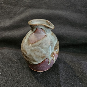 May include: A small, hand-thrown ceramic vase with a rounded body and a wide, uneven opening. The vase is glazed in shades of brown, gray, and white, with a hint of pink.