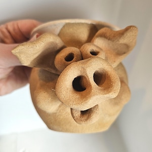 May include: A tan-colored ceramic mug shaped like a pig, featuring a prominent snout with two nostrils, two round eyes, and two large ears extending from the sides. The surface has a textured, matte finish, and the mug has an open mouth.