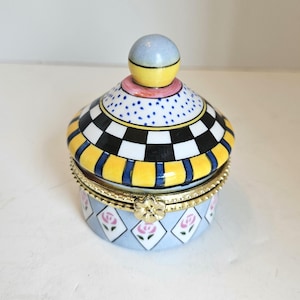 May include: A decorative trinket box with a whimsical design. The lid features a checkerboard pattern, yellow stripes, and a blue and yellow knob. The base is light blue with pink rose accents and a gold-colored trim.