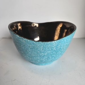 May include: A turquoise ceramic bowl with a textured exterior and a dark brown interior. The bowl has a unique, heart-shaped design, perfect for serving snacks or as a decorative piece. The bowl's dimensions are not visible in the image.