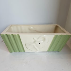 May include: A rectangular ceramic planter featuring a cream-colored center panel with a raised elephant relief design. The two side panels are finished in a light green color with vertical fluted ridges, creating a structured and clean appearance.