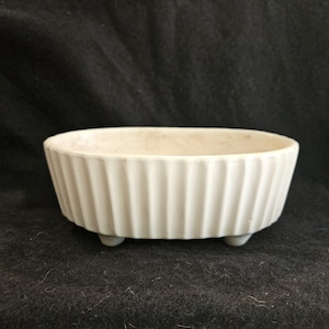 May include: A small, off-white ceramic dish with a ribbed exterior and three small feet. The oval-shaped bowl is a light cream color and sits on a dark background. The dish is likely for serving or display.