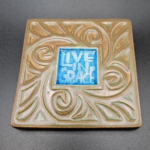 May include: Square ceramic tile with a bronze and green floral design. The center features a blue square with the words "LIVE IN GRACE" in white lettering. The tile has a textured surface.