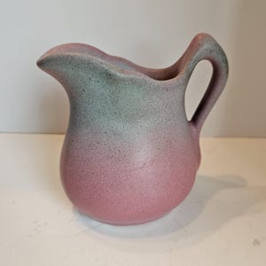 May include: A small, ceramic pitcher with a handle and a spout. The pitcher has a gradient color scheme, transitioning from a dusty rose at the base to a muted green at the rim. The surface has a speckled texture.