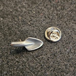 May include: A silver-toned metal pin with a shovel-shaped design. The pin has a safety clasp on the back.