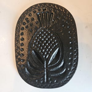 May include: Black, oval-shaped cast iron trivet with a raised thistle design. The trivet features a detailed, textured surface with a decorative border of dots and triangles. The thistle is the central focus, with intricate detailing.