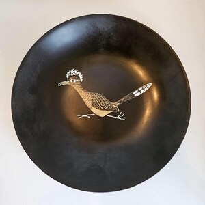 May include: A round, black decorative bowl with a roadrunner bird design. The bird is depicted in shades of brown, white, and tan, with a distinctive crest. The bowl has a smooth, glossy finish.