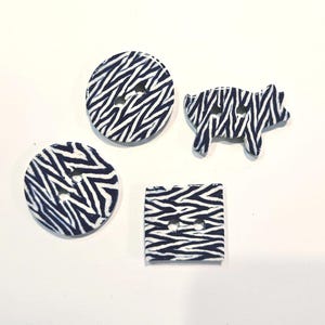 May include: Four decorative buttons with a zebra print pattern in navy blue and white. The buttons are in different shapes: two round, one square, and one pig-shaped. Each button has two holes for sewing.