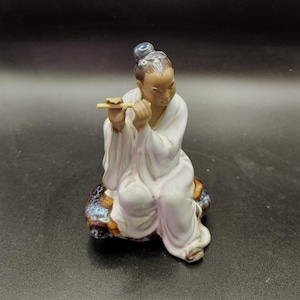 May include: A ceramic figurine of a seated figure playing a flute. The figure is dressed in a white robe and has a topknot. The figurine is seated on a rock with blue, brown, and gold accents. The background is black.