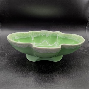 May include: A light green ceramic bowl with a scalloped, multi-lobed rim and a pedestal base. The interior and exterior edges feature a white drip glaze effect, creating a contrast against the smooth, solid light green finish of the vessel.