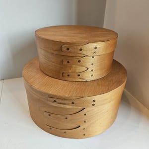 May include: Two wooden Shaker-style oval boxes, one stacked on top of the other. The boxes are made of light-colored wood and have a natural finish. Each box has a lid and is secured with wooden bands and small metal tacks.