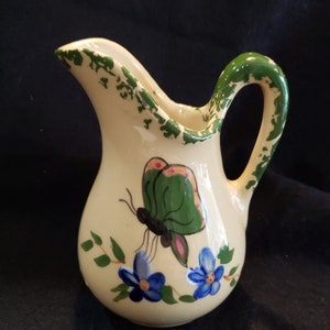 May include: A small, cream-colored ceramic creamer with a green handle and a painted butterfly design. The butterfly has green wings with pink accents and is surrounded by blue flowers and green leaves.