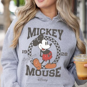 Op de afbeelding: Een grijze hoodie met een vintage Mickey Mouse-print. De print toont Mickey Mouse in een geruite cirkel met de tekst "Mickey Mouse" en "1928" boven en onder de cirkel. De tekst "Disney" staat onder de cirkel.