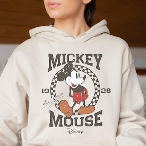 Op de afbeelding: Een cr&egrave;mekleurige hoodie met een vintage Mickey Mouse-afbeelding. De afbeelding toont Mickey Mouse in een rood-zwarte outfit, met de tekst "Mickey Mouse" en "1928" in een zwart-wit geblokte cirkel.